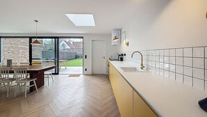 A modern kitchen with a large island, a dining area with a herringbone floor, and a view of a backyard through large windows. - Maison Martha - Oostduinkerke (Koksijde)