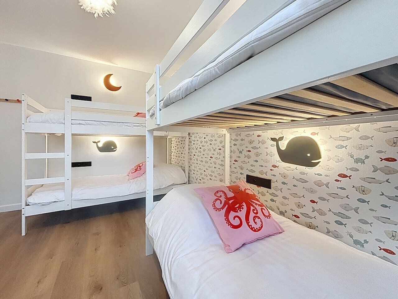 A bunk bed room with a moon-shaped light fixture above the beds, a whale decal on the wall, and a pink octopus pillow on the lower bunk.