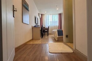 A hotel room with a wooden floor, a desk with a chair, a bed with a white cover, and a window with curtains.