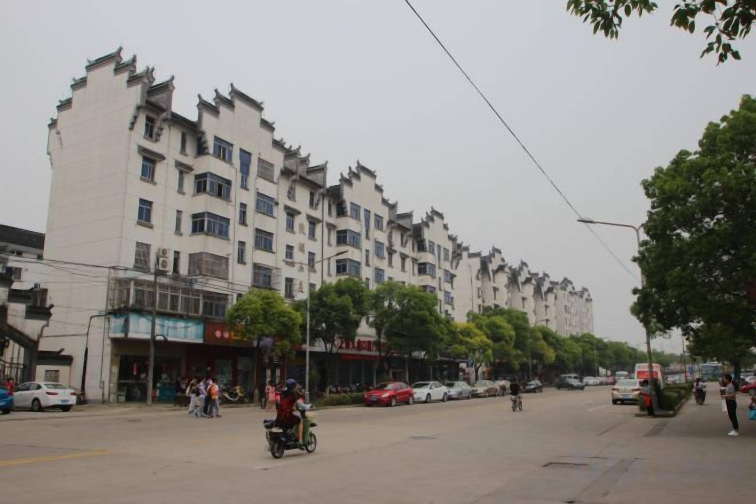 Una calle con un edificio de varios pisos, coches aparcados y algunas personas.