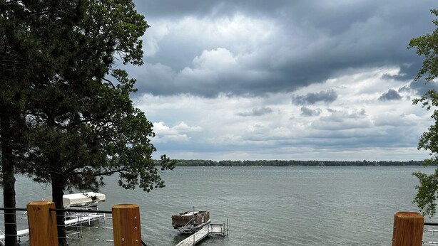 A wooden deck with a view of a lake, dock, and grassy area with tables.
