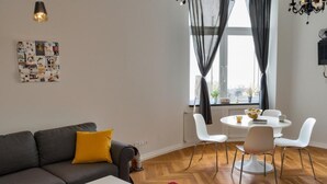 Uma sala de estar moderna com um sofá cinzento, uma mesa de centro branca e um tapete vermelho. Há uma área de jantar com uma mesa e cadeiras brancas, e uma janela com cortinas leves.