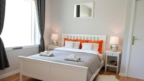 Quarto bem arrumado com cama branca, duas mesinhas de cabeceira com luminárias, um espelho na parede e um tapete macio no chão.