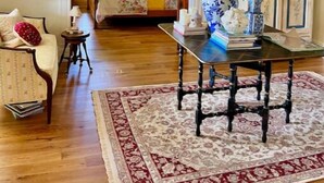 Una habitación con piso de madera, una mesa con un jarrón de flores, un sofá con un cojín rojo y una cama con colcha estampada.