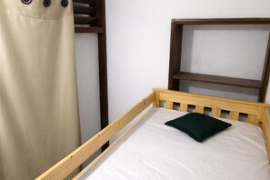 Una cama de madera individual con colchón blanco y almohada verde oscuro, cabecero de madera y cortina color beige.
