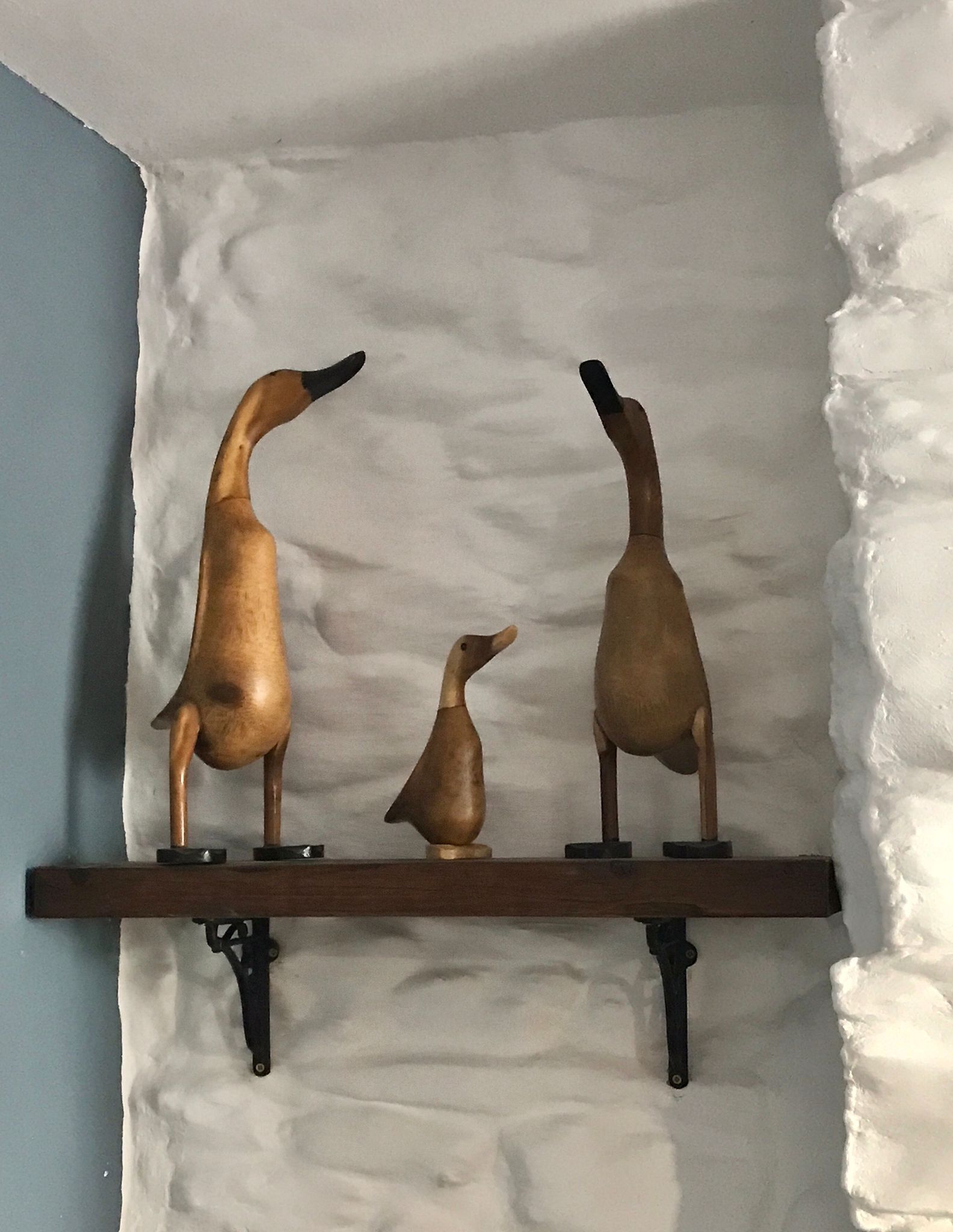 Une étagère en bois ornée de trois sculptures d’oiseaux, le tout placé devant un mur à texture.
