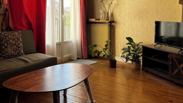 Un salon avec un plancher en bois, un canapé, une table basse, un téléviseur, un lustre et une fenêtre avec des rideaux.