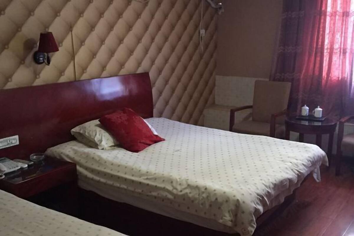 A hotel room with a tufted headboard, a bed with a red pillow, a bedside table with a phone, and a small table with two bottles.