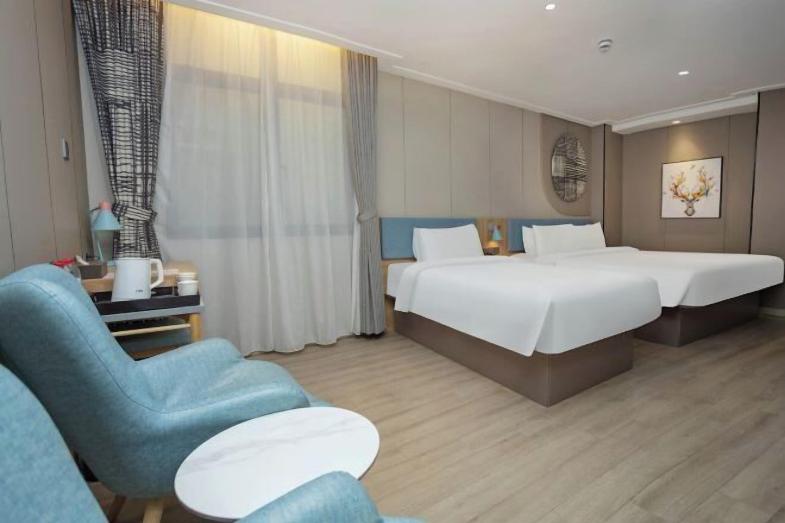A hotel room with two beds, a desk with a lamp, a chair, and a large window with curtains.