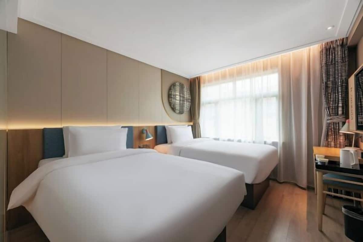 Une chambre d’hôtel avec deux lits, un bureau, une chaise, une lampe et une grande fenêtre avec des rideaux.
