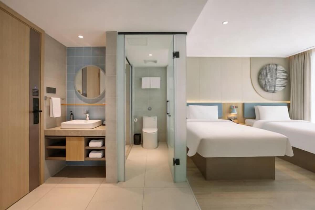 A modern hotel room with a glass partition separating the bathroom, a sink with a mirror, and two beds with white bedding.