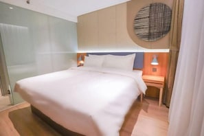 Una habitación de hotel moderna con una cama grande, una mesita de noche con lámpara, una luz circular montada en la pared y un box de ducha de vidrio.