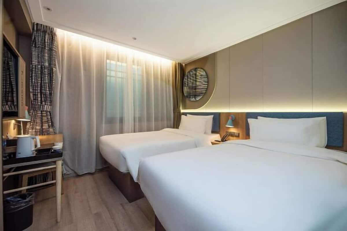 Une chambre d’hôtel avec deux lits, un bureau avec une lampe, une poubelle et une grande fenêtre avec des rideaux.