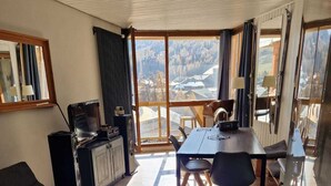 A modern living room with a sofa, coffee table, and dining area. There is a television and a cabinet. Large windows offer a view of the outdoors. - New for rent, spacious 65m² duplex facing south.  (Val d'Isère)