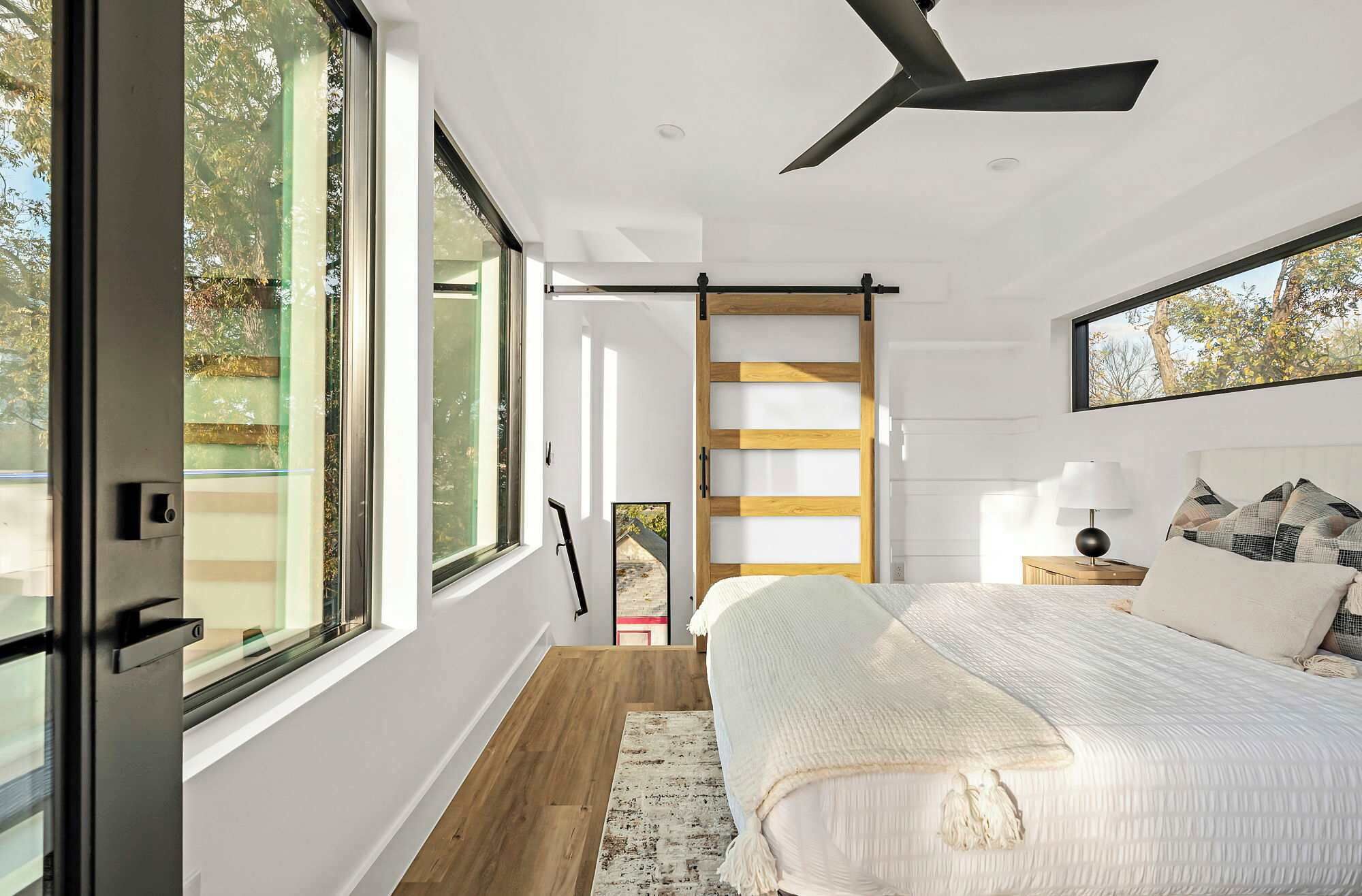 Ein Schlafzimmer mit einem Bett, einem Nachttisch, einer Lampe, einem Deckenventilator und großen Fenstern.