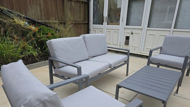 Een terras buiten met een grijze metalen tafel en twee grijze stoelen met kussens.