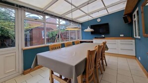 Een eethoek met een grote tafel en stoelen, een televisie aan de muur bevestigd, en uitzicht op het buitengebied door grote ramen.