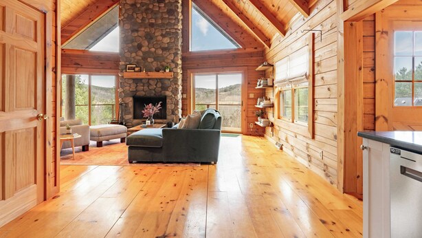 A log cabin interior with a stone fireplace, wooden floors, and large windows offering views of the outdoors. - Secluded w/ Deck Views, Hot Tub, & Ping Pong Hudson in the Berks by Avantstay (East Chatham)
