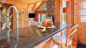 A wooden cabin interior with a kitchen area featuring a granite countertop, a stone fireplace, and a large window. - Secluded w/ Deck Views, Hot Tub, & Ping Pong Hudson in the Berks by Avantstay (East Chatham)