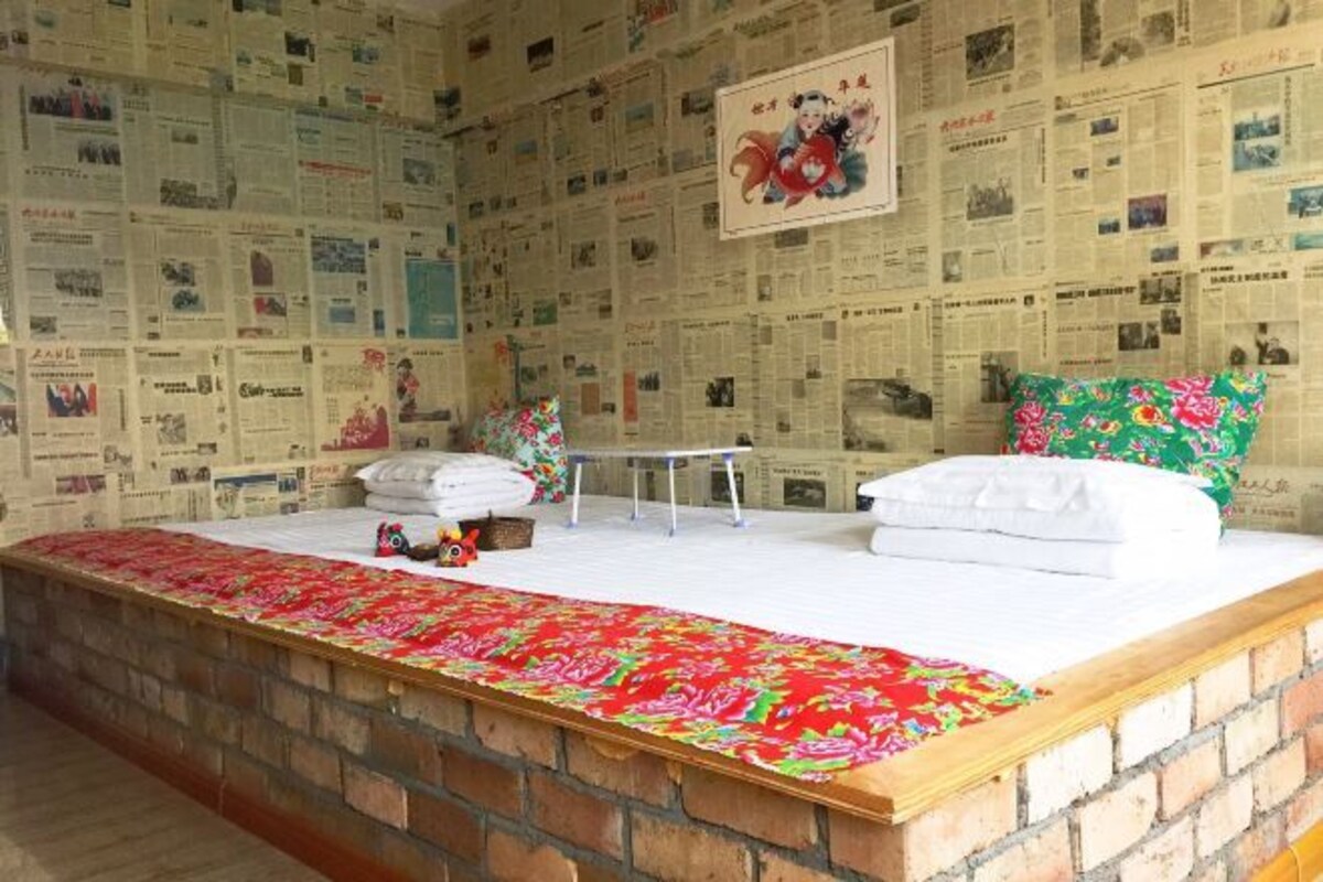 Una habitación con pared de ladrillo, cama con ropa blanca, una mesita pequeña y un cojín floral colorido.