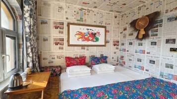 A hotel room with a bed, a wooden table, a kettle, and a wall covered with newspaper clippings.