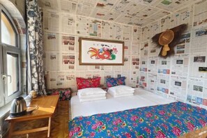 Una habitación de hotel con una cama, una mesa de madera, un hervidor y una pared cubierta de recortes de periódico.