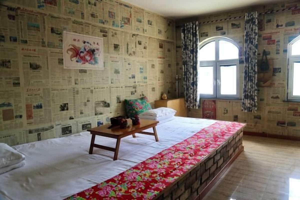 A room with a bed, a wooden nightstand, a window with curtains, and a wall covered with newspaper clippings.