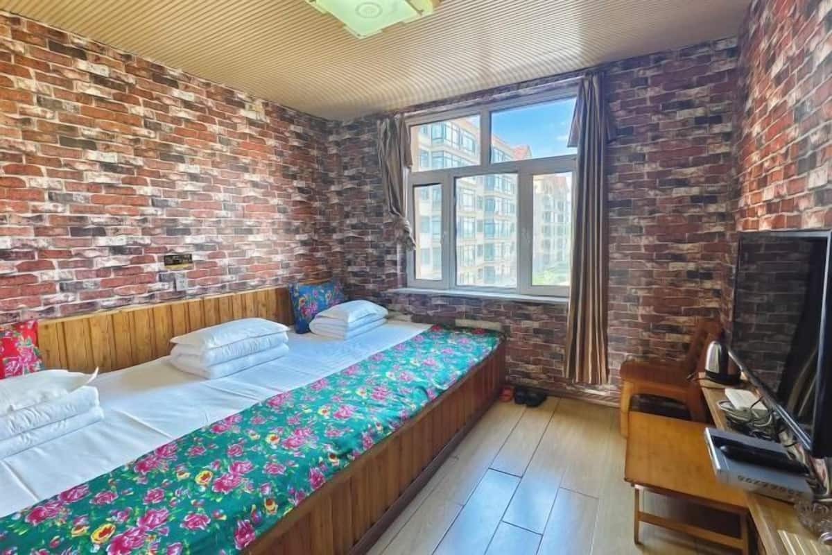A room with a brick wall, a bed with floral bedding, a window with curtains, and a TV on a wooden table.