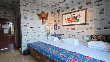 A room with a bed covered in white linens, a colorful floral bedspread, and a wall decorated with numerous small pictures.