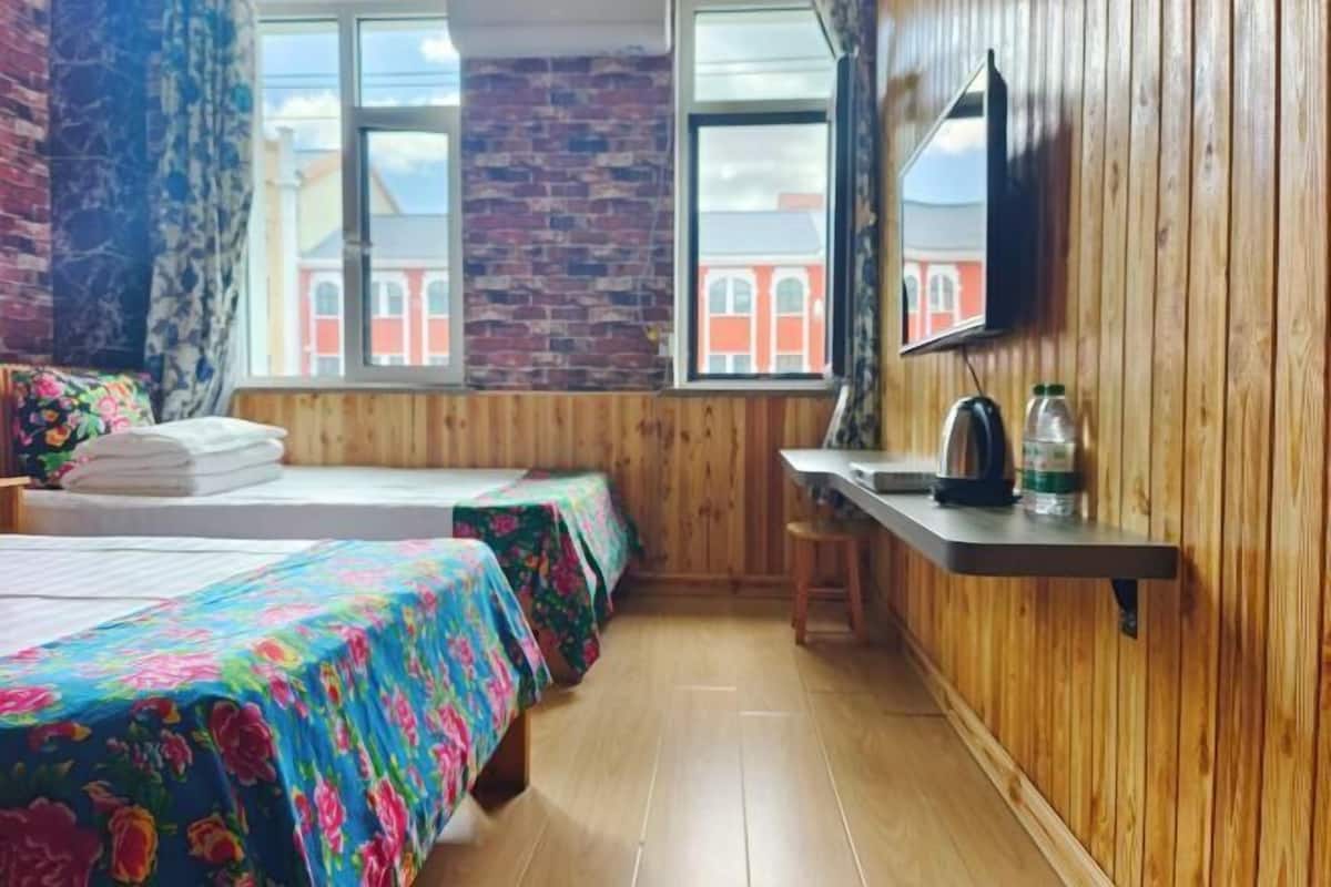 A room with wooden walls, a brick wall, two beds with floral bedspreads, a wooden desk, a chair, a TV, and a window with a view of buildings.