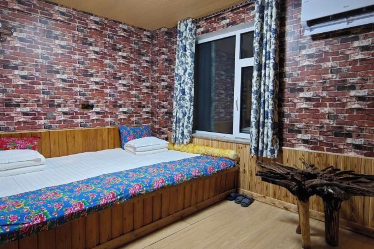 Una habitación con pared de ladrillo, cama de madera con colcha colorida, ventana con cortinas y una mesa de madera.