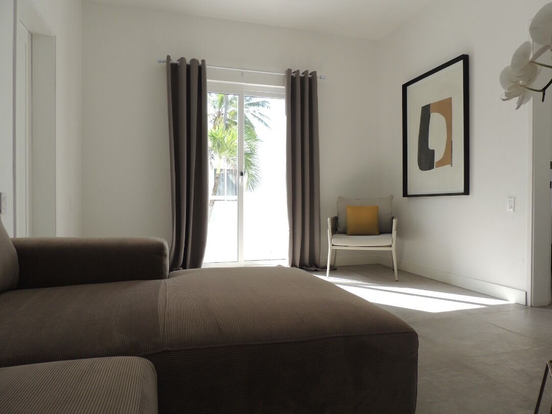 Une chambre d’hôtel moderne avec un grand lit, un fauteuil avec un coussin, une œuvre d’art encadrée au mur et une vue sur les palmiers par la fenêtre.