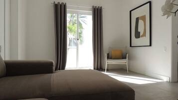 A modern hotel room with a large bed, a chair with a cushion, a framed artwork on the wall, and a view of palm trees through the window.