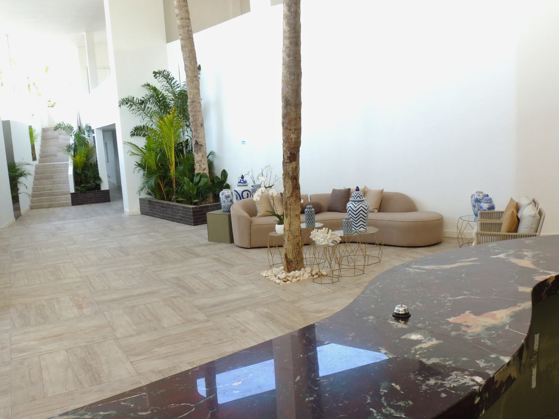 A spacious living area with a curved black marble table, a beige sofa set, and a variety of plants.