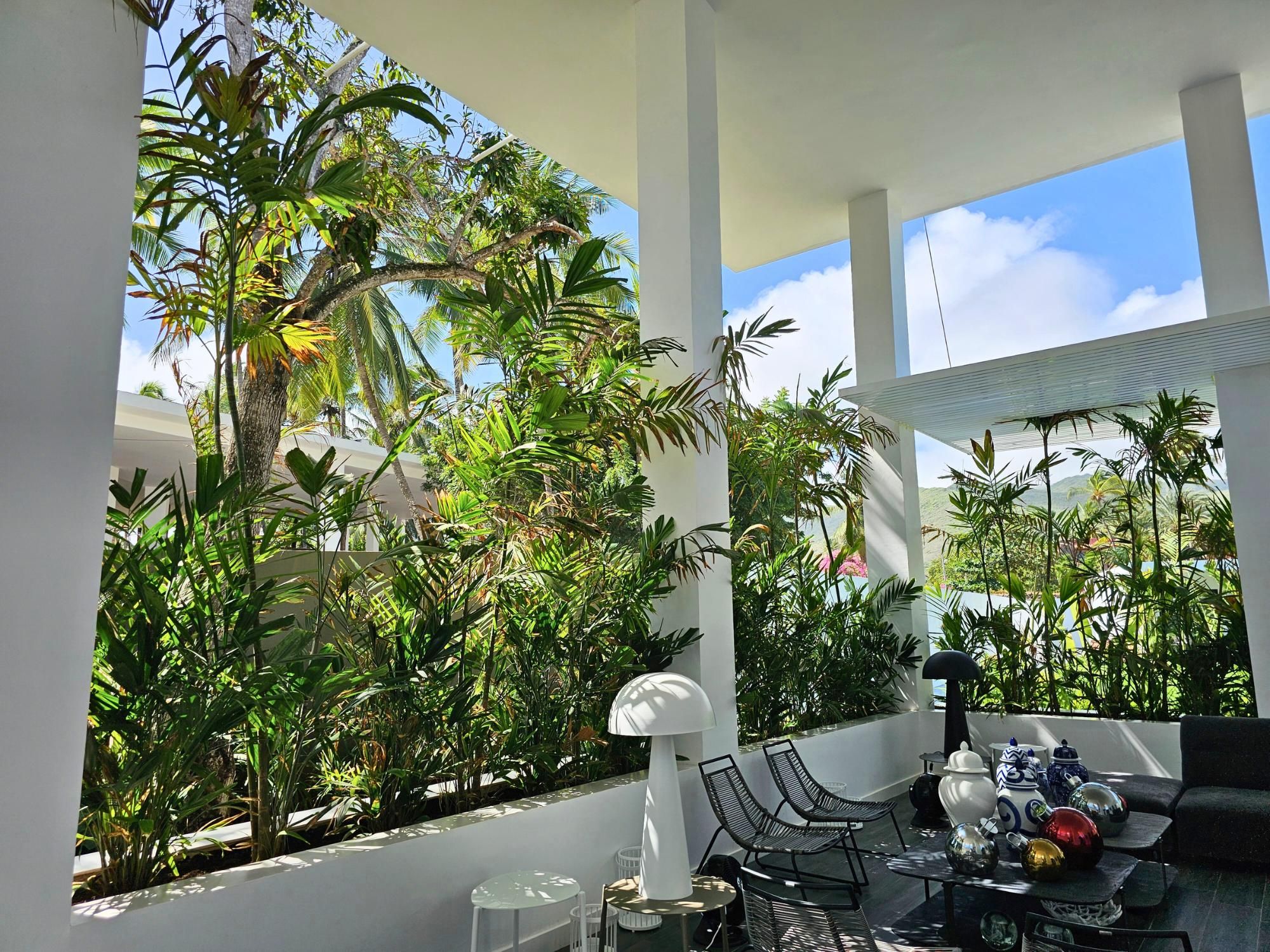 A modern outdoor seating area with a view of lush tropical vegetation, white pillars, and a clear sky.
