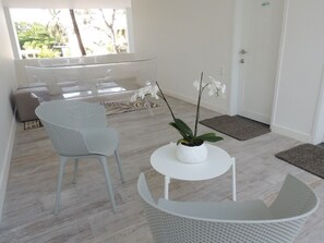 Una habitación moderna con una silla blanca, una mesita pequeña y una orquídea en maceta. Se ve una puerta y una ventana con vistas a zonas verdes.