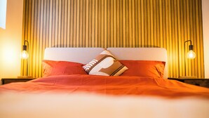 Una cama con un almohadón a rayas y una manta roja, cabecera de listones de madera y dos lámparas fijadas a la pared.