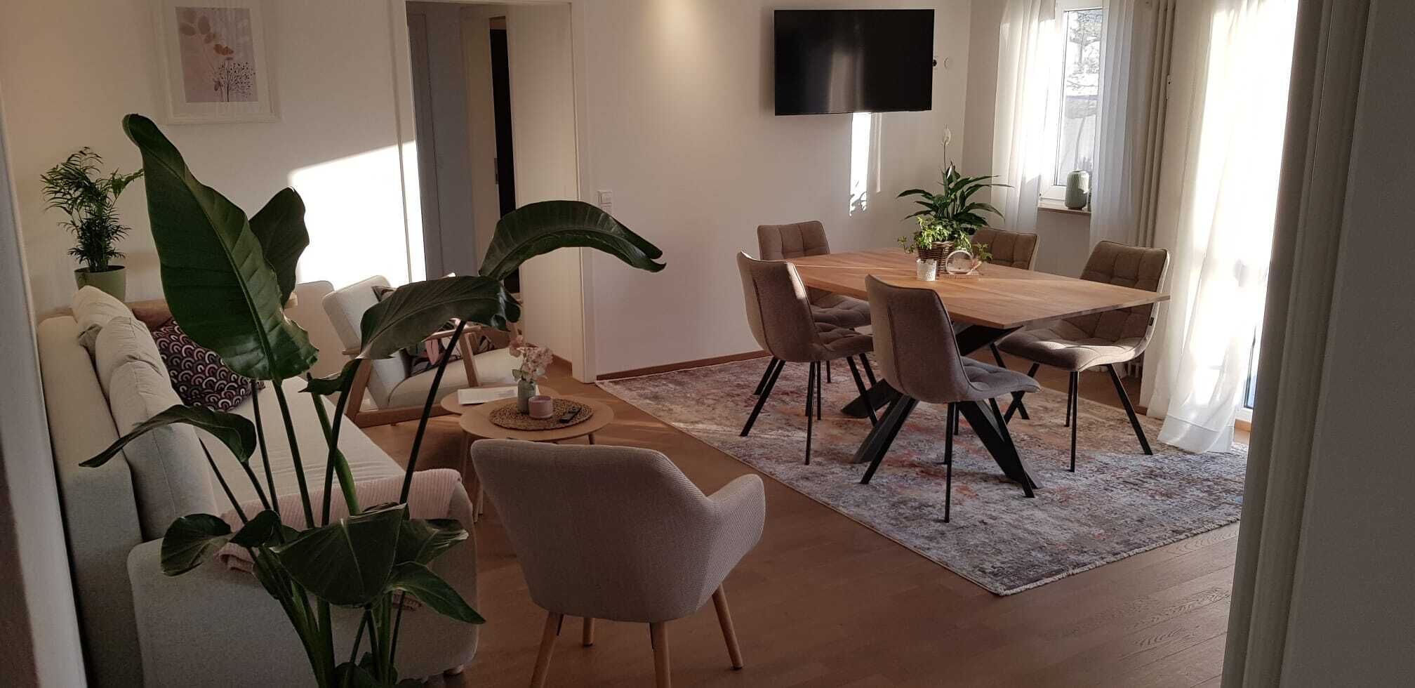A modern living room with a dining area, featuring a large wooden table, comfortable seating, and several potted plants.