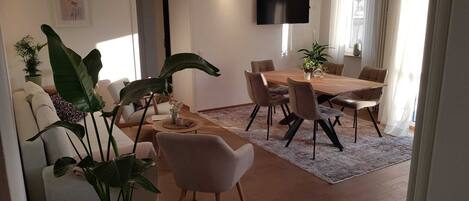 A modern living room with a dining area, featuring a large wooden table, comfortable seating, and several potted plants.