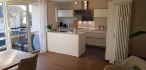 A modern kitchen with a central island, a dining area with a table and chairs, and a view through a large window.