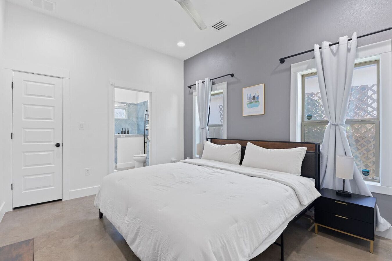 A bedroom with a bed, a nightstand, a window with curtains, a door, and a bathroom visible through an open door.