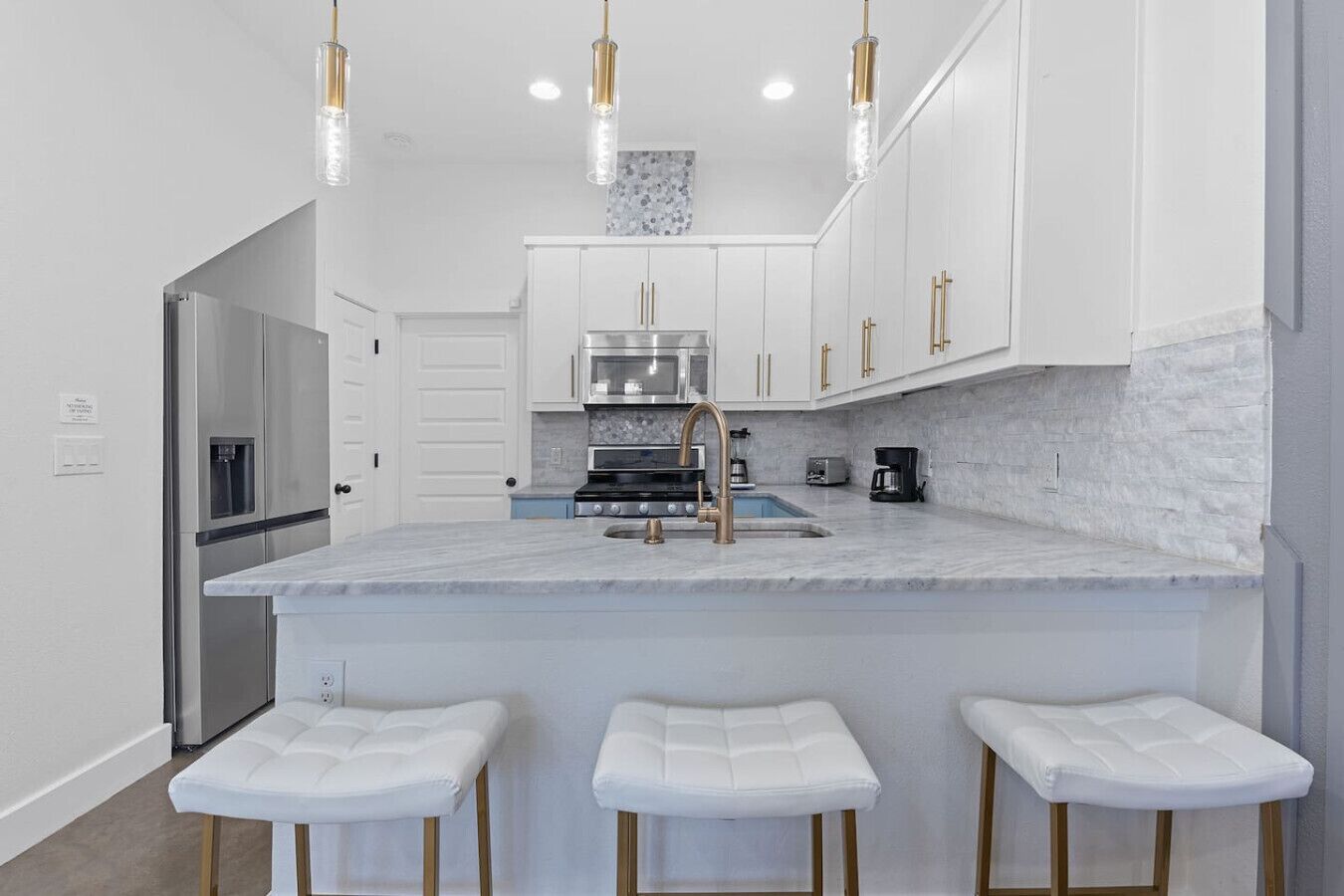 A modern kitchen with white cabinetry, a marble countertop, a stainless steel refrigerator, a built-in oven, and a sink with a gold faucet. There are three white bar stools at the island.
