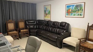 Una sala de estar con un sofá de cuero negro, sillas de madera, una mesa de centro de vidrio y dos cuadros enmarcados en la pared.