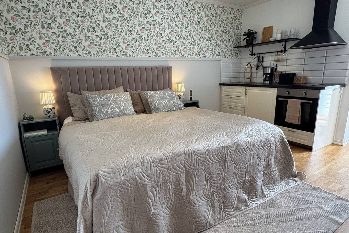 A bedroom with a large bed, a nightstand, a dresser, a built-in oven, and a kitchen area with a black range hood.