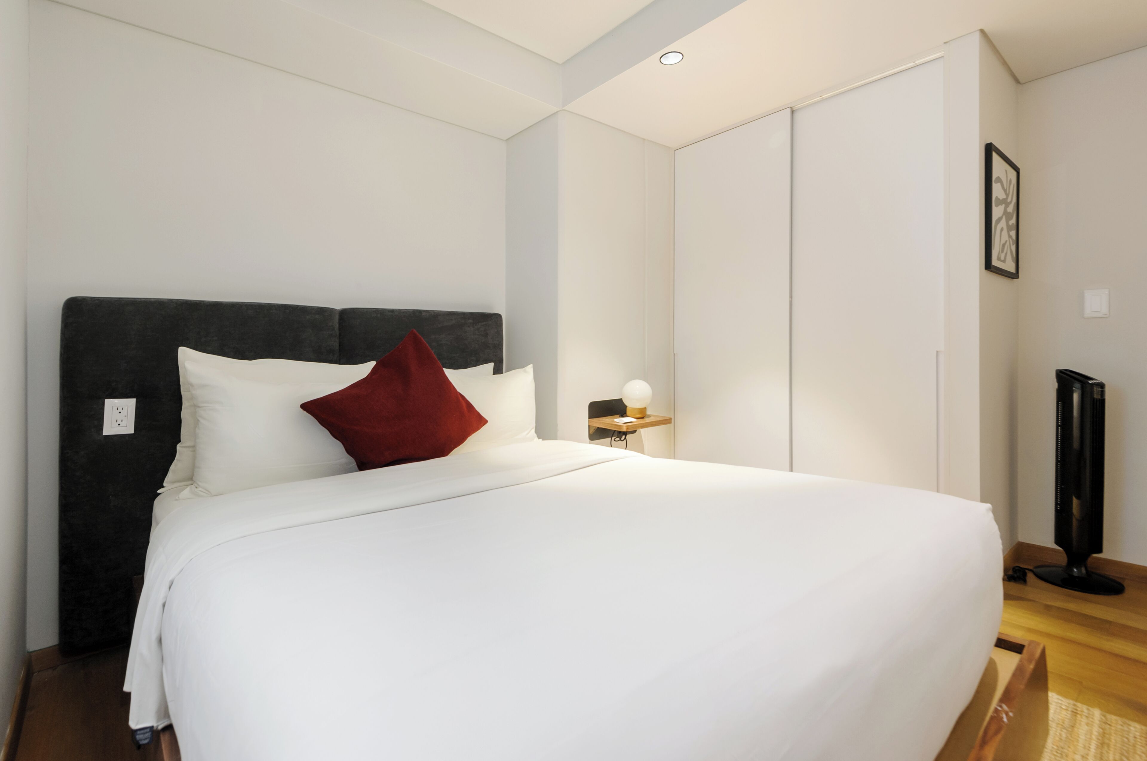 A neatly made bed with white linens and a red pillow, a bedside table with a lamp, and a framed picture on the wall.