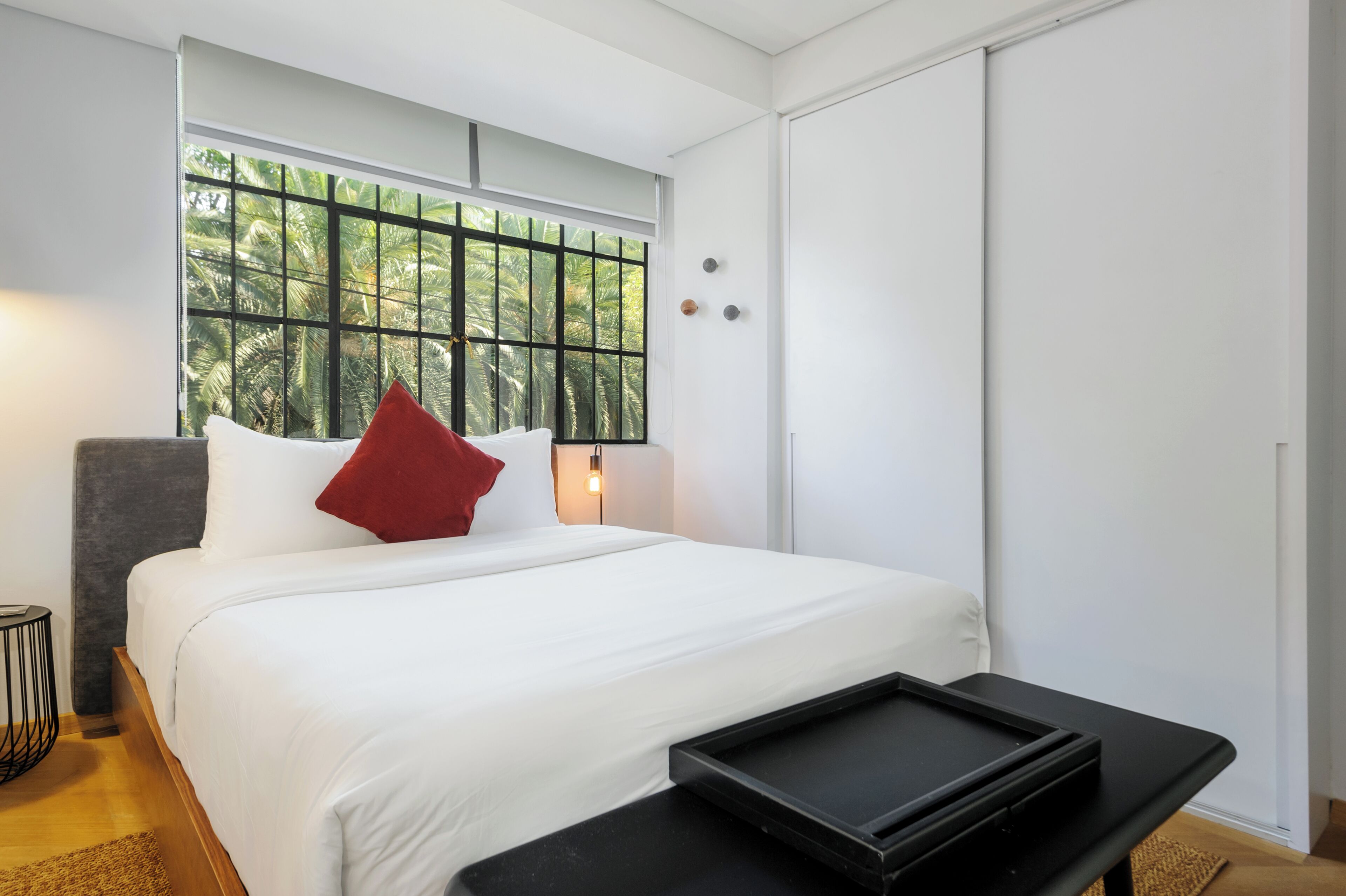 Una habitación de hotel moderna con una cama grande, una mesita de noche y un ventanal con vista a palmeras.