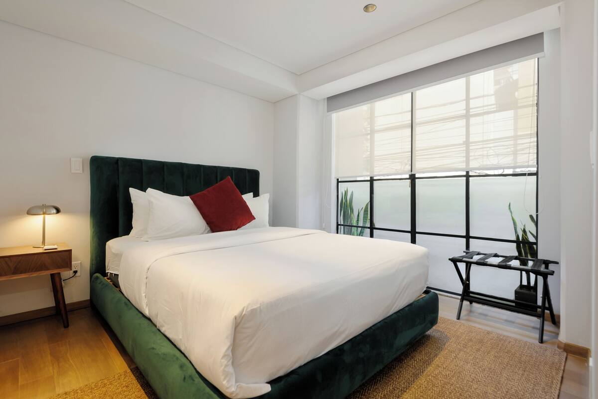 A modern bedroom with a large bed, a bedside table with a lamp, and a view of the outside through a large window.