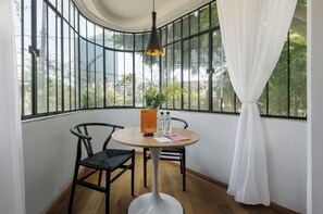 A corner room with a round table, two chairs, a hanging light fixture, a window with a view of greenery, and a curtain. - Oaxaca by Wynwood House (Mexico City)