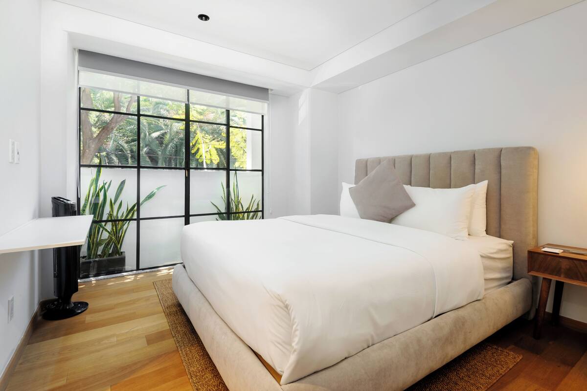 A modern bedroom with a large bed, a desk, and a window with a view of greenery.