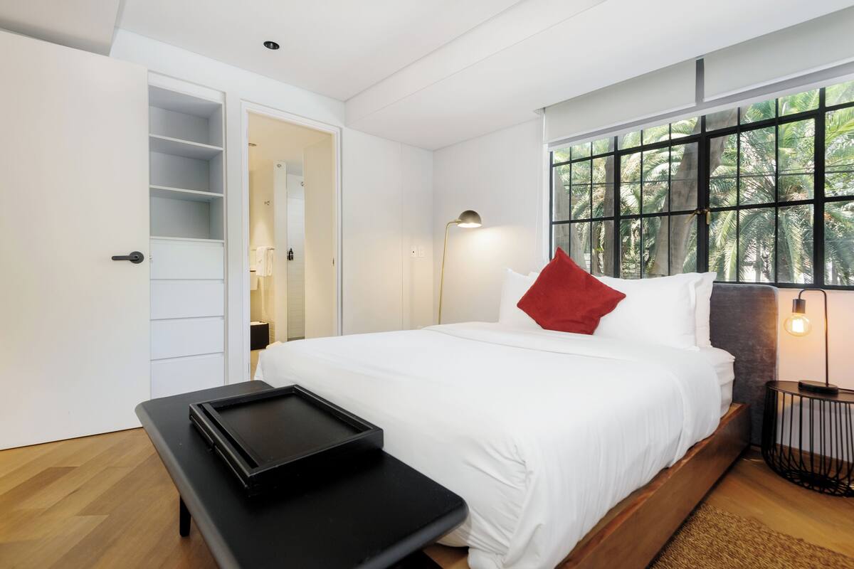 A bedroom with a bed, a bench, a bedside table, a lamp, and a window with a view of palm trees.